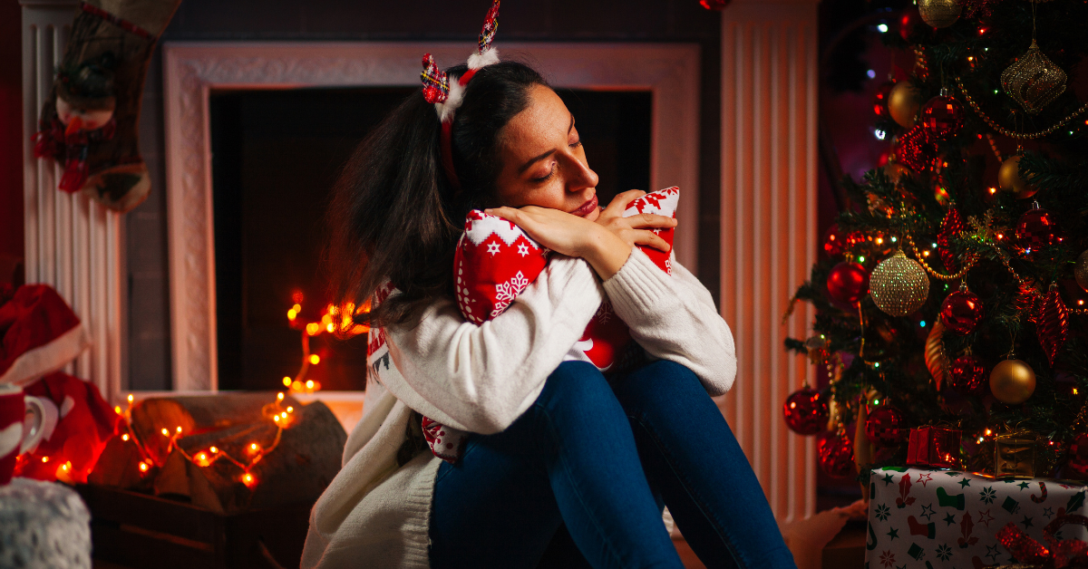 woman in holiday attire hugging red pillow in front of fireplace