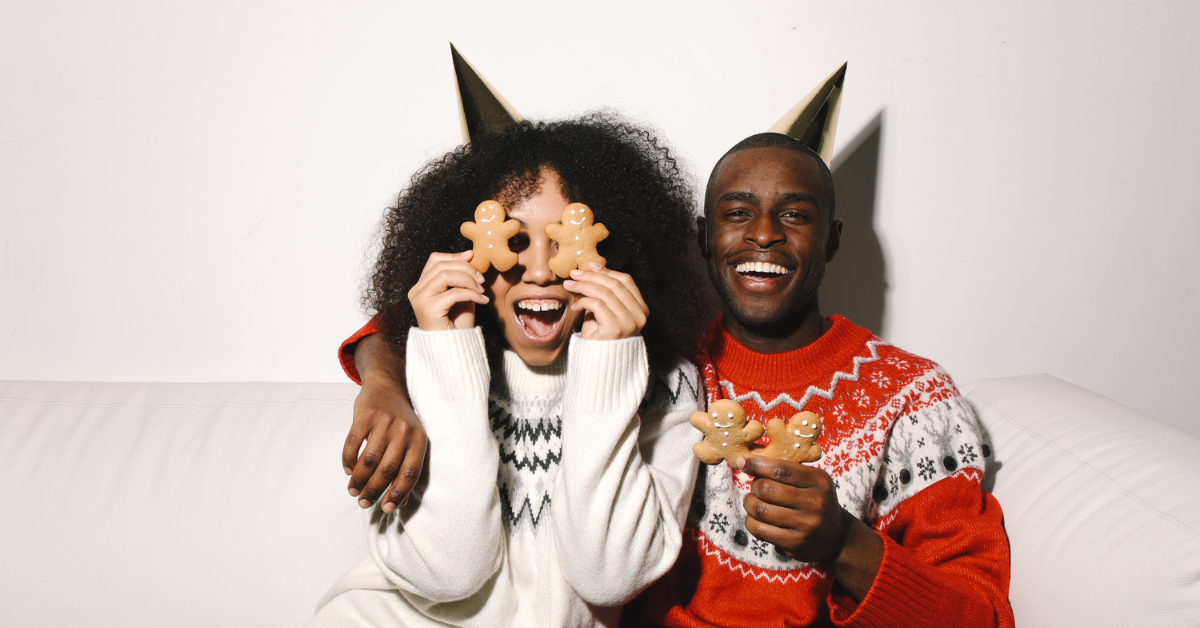 couple in holiday sweaters holding up gingerbread cookies in silly pose